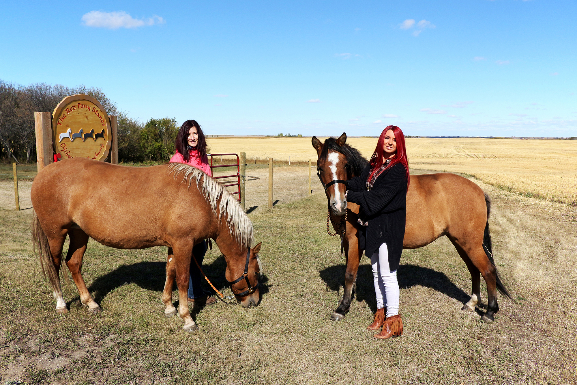 Two women stand with two horses in front of The Red Pony Stands® entrance sign
